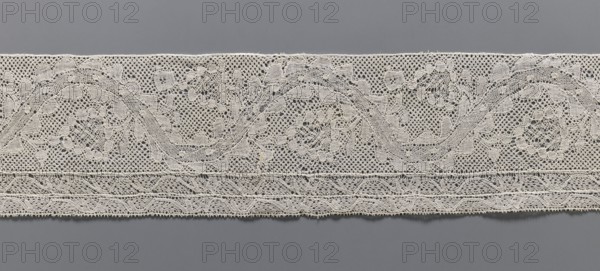 Lace fragment, bobbin lace Malines/Mechelen, 18th century; plant ornaments with floral branches and tendrils in the bends of a ribbon; straight edges, bottom edge with two trails repeating the tendril motif schematic; various mesh backgrounds; side edges folded and hemmed; width 11 cm, length 72.5 cm., A long strip of lace with intricate openwork laid horizontally against a smooth background; the lace appears off-white or cream and shows a repeating pattern of swirling floral and foliate motifs created from fine threads and tiny mesh areas, with a denser decorative band near the lower edge featuring braided and scalloped shapes; the background is a uniform gray that contrasts with the lighter lace, and the overall image emphasizes the texture and delicate holes of the fabric.