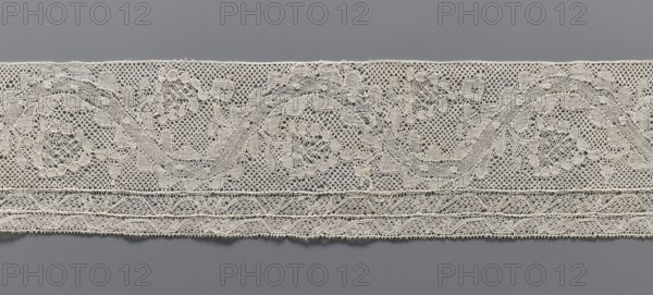 Lace fragment, bobbin lace Malines/Mechelen, 18th century. Plant ornaments: floral branches grow in the bends of a ribbon with tendrils; straight edges; bottom edge has two trails repeating the tendril motif schematically. Various mesh backgrounds. Side edges folded and hemmed. Dimensions: width 11 cm, length 72.5 cm., A horizontal strip of delicate lace in off-white or cream stretches across a neutral gray background, featuring an intricate openwork pattern with floral and leafy motifs, a few curved vine-like shapes, and several small circular voids within the mesh; the lower edge shows layered bands of denser lacework with repeating geometric scalloped details, while the texture throughout appears fine and threadlike with subtle variations in thickness and occasional tiny irregularities.