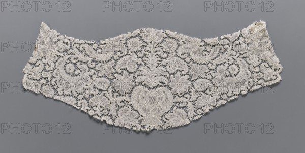 Fragment of lace, plant ornaments, bobbin lace, 18th century, Flanders, width 17 cm, length 46 cm, A delicate lace fabric piece rests flat against a gray background, showing an intricate openwork pattern of floral and foliate motifs with scalloped edges. The lace appears off-white to cream in color, with a few areas of slightly darker discoloration near one edge. The design includes several stylized flowers, curved leaf shapes, and swirling paisley-like forms, all rendered in fine thread with textured raised outlines and mesh-filled interiors. The fabric has a slightly irregular outer contour with frayed thread ends and tiny gaps where the lacework is broken, and the overall appearance is light, airy, and detailed against the solid gray surface.