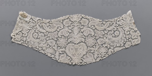 Fragment of lace, plant ornaments, bobbin lace, 18th century, Flanders, lace, width 17 cm, length 46 cm, A wide, curved piece of delicate openwork lace with an intricate floral and scrolling vine pattern, displayed against a solid background; the lace appears in off-white and cream tones and the background is gray.