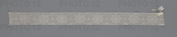 Fragment of lace, bobbin lace, 18th century, Flanders, lace, width 9 cm, length 51 cm, A long narrow strip of delicate lace laid flat against a grey background, showing an openwork pattern with a repeating floral motif and a fine net-like ground; the lace is off-white to cream in color, with subtle variations in tone along its length; at one end there are a few small attached tags and a short length of green thread visible.