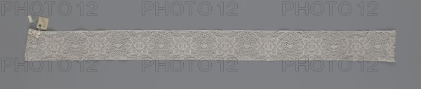 Fragment of bobbin lace, 18th century, Flanders; width 9 cm, length 51 cm; subject: lace; maker, provenance includes repossession; place: Henryków German: Heinrichau., A long narrow strip of openwork lace laid flat against a solid muted gray background, the lace appearing off-white to cream with a delicate, airy texture and an intricate repeating floral-like motif visible across its length, surrounded by a fine netted ground; one end of the lace has a small cluster of tags attached with thin strings in pale beige and light green hues, and the lace edges are slightly irregular with subtle scalloping and fine threadwork.