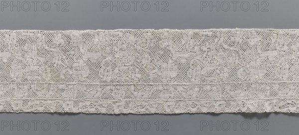 Lace fragment, bobbin lace, Binche lace, 18th century. Background mesh with large rounded meshes; edges of motifs not contoured. Large narrow laces sewn on at the bottom edge — the wider one with a motif of flower branches arranged in a twig, the narrower with a motif of a flower tendril. From Binche; width 12 cm, length 61 cm., A long strip of lace with an intricate openwork pattern displayed against a smooth background, showing a repeating floral and curvilinear design and a scalloped lower edge; the lace appears off-white or cream with subtle shading from the weave, and the background is a muted gray.