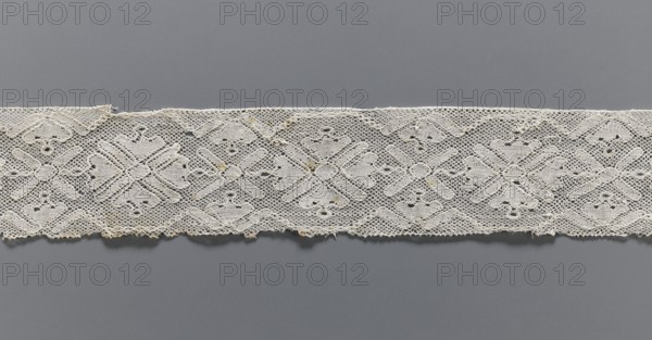Lace fragment, bobbin lace with rosette ornaments framed by a tendril motif at top and bottom; mesh background similar to a drochel; motif edges contoured and straight. 18th–19th century, Flanders. Lace, width 8 cm, length 97.5 cm., A horizontal strip of lace with a repeating geometric floral pattern spans the width of the image, placed against a smooth background. The lace appears off-white to cream with small areas of darker discoloration, and the background shows shades of gray ranging from medium to darker gray. The lace has an open, net-like texture with solid embroidered motifs forming diamond and cross shapes, and the lower edge is slightly irregular with tiny loose threads. The overall scene shows the contrast between the delicate light-colored lace and the uniform gray backdrop.