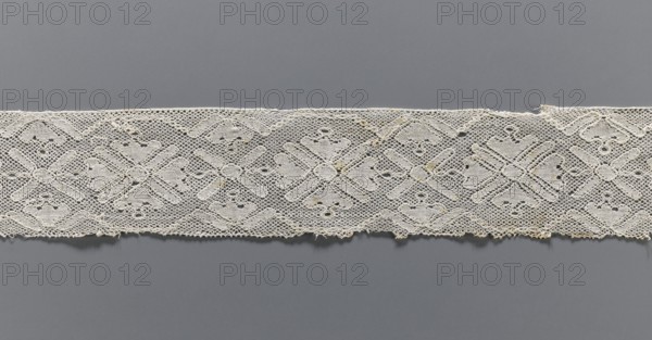 Lace fragment, bobbin lace with rosettes, the rosettes are framed by a tendril motif at the top and bottom. The mesh background is similar to a drochel. The edges of the motifs are contoured and straight. 18th/19th century, Flanders, lace, width 8 cm, length 97.5 cm., A horizontal strip of intricate lace with a repeating geometric floral-like pattern lies against a solid background; the lace is off-white or cream with areas of slight yellowing and frayed edges, featuring openwork hexagonal mesh and denser embroidered motifs that form diamond and cross shapes, and the background is a smooth medium gray.
