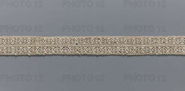 Fragment of lace, needle lace, 16th/17th century, Flanders; width 2.8 cm, length 55 cm;, A horizontal strip of delicate lace runs across the middle of the image against a solid background; the lace is light beige with an openwork floral and geometric pattern and slightly irregular edges, showing a few small loose threads and tiny darker spots, while the background is a smooth, even slate gray.
