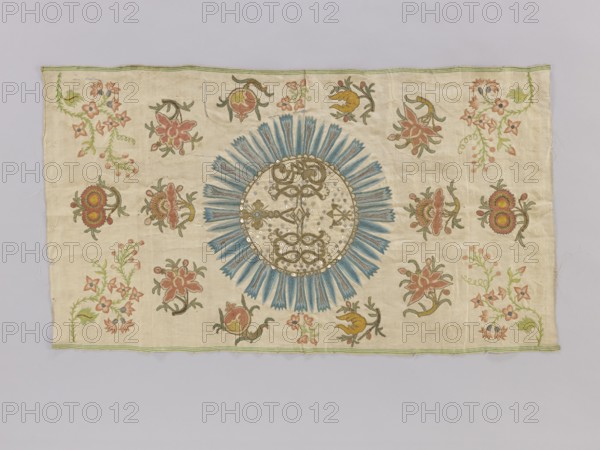 Antependium, antependium; fragment of church embroidery on cream silk satin 46 gauge, green selvedges with embroidery and glued appliqué of colorful silk threads, gold and silver threads, and sequins. In the center, a large radiant medallion bears the IHS monogram with a cross and three nails. Background filled with projections of stylized floral sprigs; small sprigs in the corners and on the sides of the medallion are embroidered, remaining background motifs are appliquéd glued. 18th century, Poland; fabric; height 82 cm, width 47 cm., A rectangular piece of fabric with a light beige background decorated with a large circular sunburst motif near the center featuring blue radiating petal shapes and a circular center with a mix of beige and gold tones and reflective sequins; surrounding the central motif are a group of floral and botanical motifs in shades of pink, muted red, green, olive, yellow, and brown arranged across the fabric; the fabric edges have a narrow green trim and faint ...