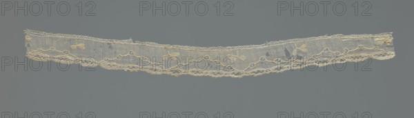 Lace fragment, needle-stitched lace, highly elongated openwork ovals. Above the edge ornament, against a mesh background, a winding line interrupted by openwork circles. In the bends of the line, a rather large three-leafed branch projection. 18th century, possibly France. Lace, width 3.5 cm, length 42.5 cm. Creator, provenance noted as repossession., A narrow strip of delicate lace in off-white and cream tones stretches horizontally across a muted gray background, featuring a fine netted ground with a scalloped lower edge and a straighter upper edge, decorative floral motifs and small leaf-like shapes worked into the fabric, a sinuous openwork border with looping patterns along the bottom, a few small holes and frayed areas, and subtle variations in the off-white and beige threads against the gray backdrop.