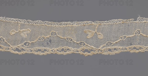 Lace fragment, needle-stitched lace, highly elongated openwork ovals. Above the edge ornament, against a mesh background, a winding line interrupted by openwork circles. In the bends of the line, a rather large three-leafed branch projection. 18th century, probably France. Width 3.5 cm, length 42.5 cm., A horizontal length of delicate lace in a creamy off-white color stretches across a gray background, featuring a fine hexagonal mesh ground with a scalloped lower edge and a looser decorative upper edge; the lace shows a repeating floral motif with small three-petaled leaf shapes and a sinuous vine of linked rings and oval loops, plus a few irregular open areas and frayed thread ends.