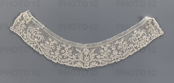 Collar, 19th century, France, fashion accessory—lace collar with floral ornaments. Band of equal width, arched; outer edge slightly serrated following the ornament forms. Ornament placed on a mesh background. Width 5.2 cm, length 37 cm., A curved lace collar displayed against a solid gray background, featuring an off-white delicate net base with ornate floral and scrollwork embroidery along its lower edge and a scalloped border; a few small fabric tags are attached near one end.