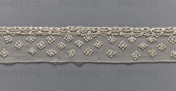 Fragment of lace, needle lace, Alençon lace, 19th century, France, Henryków German: Heinrichau, lace, width 6 cm, length 93 cm, A horizontal strip of delicate lace in cream color is centered against a smooth gray background, featuring a scalloped ornate top edge with looping floral motifs and a straight bottom edge with slight irregularities; the lace body has a fine hexagonal net pattern decorated with a repeating arrangement of small clustered floral rosettes in cream, creating a rhythmic pattern across the strip.