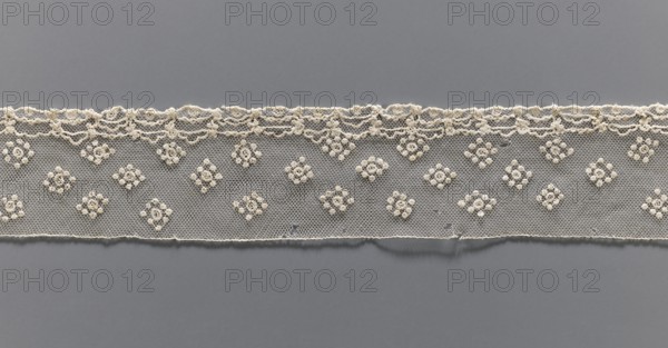 Fragment of lace, needle lace Alençon lace, 19th century, France, from Henryków German: Heinrichau, lace, width 6 cm, length 93 cm, A horizontal strip of delicate off-white lace runs across a gray background, showing a scalloped upper edge with a textured trim and a straight lower edge. The lace has a fine hexagonal netting with a repeating pattern of small floral-like clusters raised in off-white thread, spaced evenly across the strip. The scalloped top features loops and small rounded motifs in the same off-white color, and there are subtle shadows where the lace lies against the flat gray surface. The visible colors are off-white and gray.