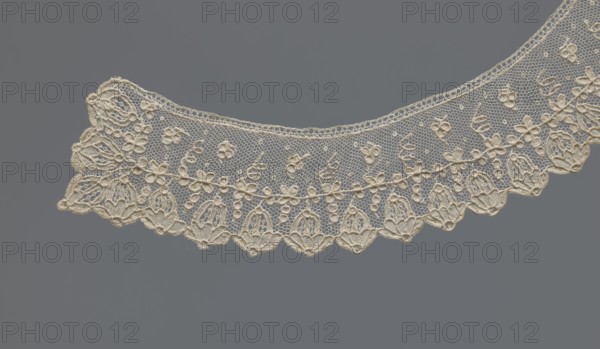 Collar, 19th century, France, band-style arched collar of equal width. Floral ornament set against a mesh background; rim with a row of flower calyxes branching from a continuous branch decorated with small quatrefoils. Women's clothing accessory, lace; width 3.5 cm, length 46 cm., A delicate piece of lacework in a creamy off-white color curves across a flat background in a muted gray color, showing an open net-like ground with a scalloped lower edge and an upper edge that follows a gentle arc, featuring a repeating floral motif of small raised flowers and leafy elements connected by fine embroidered outlines, with denser embroidered sections along the scallops that form petal-like shapes and looped decorative outlines, and the overall texture appearing intricate and airy against the smooth gray backdrop.