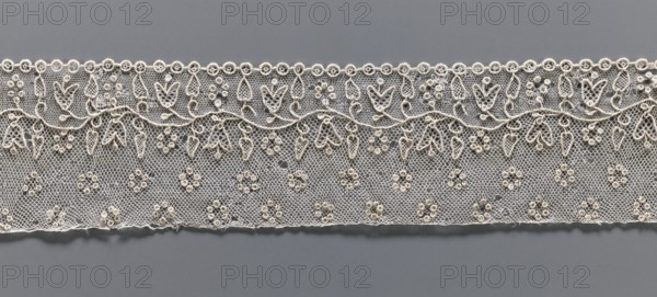 Lace fragment, needle Alençon lace; a flower branch parallel to the edge. Above the branch, in the projection, circles of small circles. Below the branch, on the edge, a row of small circles, the edge corresponding to the circles with small semicircular serrations. 18th century, France; lace; width 6.5 cm, length 33 cm., A horizontal strip of delicate lace in off-white and cream tones lies against a smooth gray background; the lace features a mesh net base with a repeating floral and leaf pattern made from raised embroidery, including a scalloped top edge with small circular motifs, a sinuous vine with teardrop and heart-shaped leaves, and a field of small clustered rosettes, with visible texture variations where threads loop and knot and some slight irregularities along the lower edge.