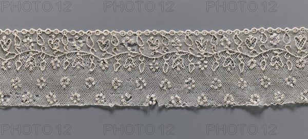 Lace fragment, needle lace Alençon, 18th century, France;, Description: a flower branch parallel to the edge; above the branch, in the projection, circles made of small circles; below the branch, on the edge, a row of small circles with the edge corresponding to the circles—small semicircular serrations. Dimensions: width 6.5 cm, length 32 cm. Repossession, A horizontal strip of delicate lace with an intricate floral and looped pattern sits against a smooth background; the lace is predominantly off-white or cream with a fine netting base and raised embroidered motifs that include a scalloped top edge of small circular loops, a sinuous vine-like band, and a repeated arrangement of teardrop and petal shapes dangling above a field dotted with small clustered rosettes; the background visible through the lace and surrounding it is a uniform medium gray, and the overall image shows subtle shadows and slight irregularities along the lower edge of the lace.