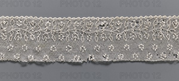 Lace fragment, needle lace Alençon lace, A flower branch parallel to the edge. Above the branch, in the projection, circles made of small circles. Below the branch, on the edge, a row of small circles, the edge corresponding to the circles—small semicircular serrations. 18th century, France, lace, width 6.5 cm, length 32 cm, A horizontal strip of delicate lace in shades of off-white and cream lies against a flat gray background, showing a repeating floral and geometric pattern with a scalloped top edge and a straighter bottom edge; the lace fabric has a fine hexagonal mesh with a few raised knot-like floral motifs and looping vine-like designs, and there are small irregular frayed threads along the lower edge.