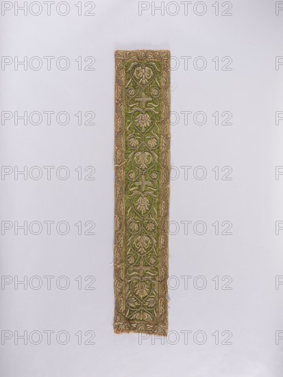 Chasuble column, embroidery on light green satin fabric bordered with pink-salmon fabric; embroidered with a pattern of interconnected heart-shaped palmettes with flowers in the center, small crosses between the palmettes, and a border with a simplified leafy tendril pattern. Embroidery: raised, on a hem, on a cord backing, with gold-braided metal thread and gold sequins. Liturgical vestment / church parament used in churches and chapels; plant ornaments, sacred art. 17th/18th century, Poland. Materials: fabrics; dimensions: length 96 cm, width 18.5 cm., A narrow vertical fabric strip centered on a pale background, showing an ornate repeating floral and vine pattern. The central field is green with a raised design in a metallic beige or gold color, and the border is a matching metallic beige or gold with intertwined motifs. The fabric shows some frayed edges and loose threads and the green area has textured, slightly worn patches. Overall colors visible are green, beige or gold, and th...