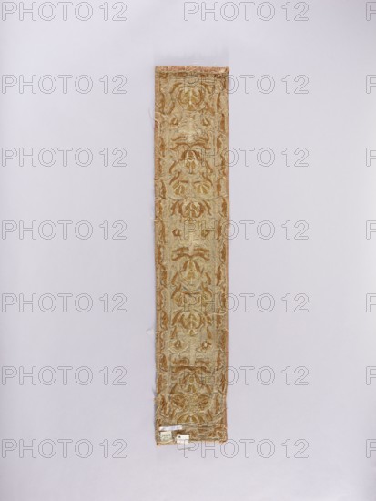 Chasuble column, embroidery, church paraments; flowers, leaves, liturgical vestments, plant ornaments, sacred art, churches and chapels furnishings. Chasuble column made of light green satin fabric, bordered with pink-salmon fabric, embroidered with a pattern of interconnected heart-shaped palmettes with flowers in the center. Small crosses between the palmettes. Border with a simplified leafy tendril pattern. Embroidery: raised, on a hem, on a cord backing, with gold-braided metal thread and gold sequins. 17th/18th century, Poland. Fabrics; length 96 cm, width 18.5 cm., A narrow rectangular textile strip with ornate repeating floral and foliate patterns in shades of gold and beige, mounted vertically against a pale background; the strip shows a decorative border and slightly frayed edges with loose threads, and small tags attached near one end, with overall colors including gold, beige, brown, and hints of off-white.