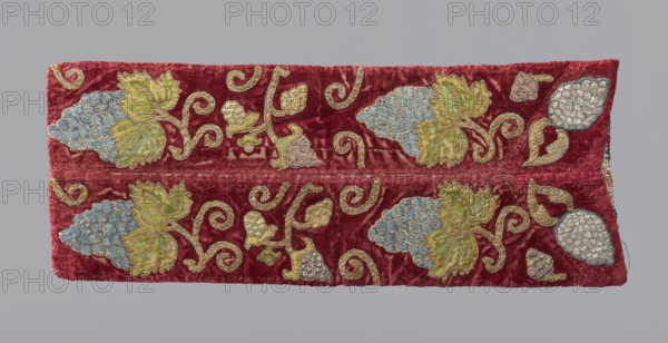 Fragment of a chasuble column, embroidery, fragment of a larger textile object, vines, liturgical vestment. Triangular cutout and traces of ripped braids visible at the top. Two strips of red velvet sewn together, embroidered with large and smaller grape vines. Flat embroidery in blue and green silk thread and gold and silver metal thread. Ornament arrangement indicates fabric comes from a larger piece. Reverse: wool embroidery on canvas with geometric and floral motifs in green, red, and black. 18th century, Poland. Fabrics; length 46 cm, width 18 cm., A rectangular textile panel with a rich red background featuring an embroidered repeating vine motif; the embroidery includes leafy shapes in shades of green and yellow, clustered scale-like elements in pale blue and off-white, and swirling stems and outlines in a metallic gold tone, with the red velvet showing variations in texture and sheen against a neutral gray backdrop.