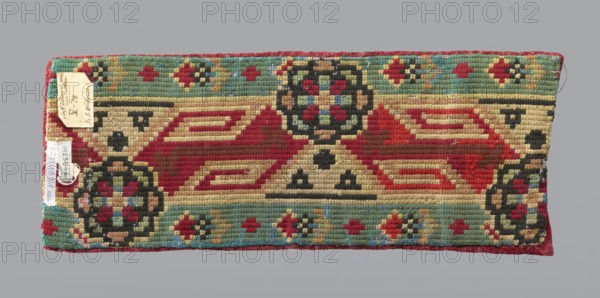 Fragment of a chasuble column, embroidery, fragment of a larger textile object, vines, liturgical vestment. Triangular cutout and traces of ripped braids visible at the top. Two strips of red velvet sewn together, embroidered with large and smaller grape vines. Flat embroidery in blue and green silk thread and gold and silver metal thread. Ornament arrangement indicates the fabric derives from a larger piece. Reverse: wool embroidery on canvas with geometric and floral motifs in green, red, and black. 18th century, Poland. Fabrics; length 46 cm, width 18 cm., A rectangular woven textile with a patterned design featuring geometric motifs and floral-like roundels in a palette of red, green, beige, black, turquoise, blue, and touches of brown, edged with a red border; the central area shows large red diamond shapes on a beige background with black triangular accents, flanked by rows of turquoise bands containing small floral motifs in red, green, blue, beige, and brown, and larger circula...