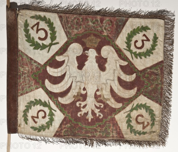 Double-sided scout pennant with painted decoration and inscriptions; vexil featuring a crown motif, Scout cross, and White Eagle coat of arms; painted fabrics; maker, origin/provenance, before 1939; Poland, Warsaw; height 53 cm, width 63.5 cm., A rectangular textile flag with fringed edges, mounted on a vertical wooden staff at the left edge, showing a central stylized bird motif in a lighter off-white color against a reddish-brown background, surrounded by scalloped shapes in the same light color and outlined with a zigzag green border; the corners contain light diamond panels each with a green laurel wreath enclosing a curved dark mark, and the surrounding areas show a mottled pattern in reddish-brown and green; visible colors include off-white, reddish-brown, green, dark brown, and tan, and the fabric shows signs of wear and fading.