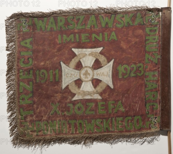 Double-sided scout pennant with painted decoration and inscriptions, before 1939; materials: painted fabrics; features: vexil, crown iconography, Scout cross, White Eagle coat of arms; origin: Poland, Warsaw; dimensions: height 53 cm, width 63.5 cm;, A square fabric banner with a worn reddish-brown field and a fringed edge in a muted brown tone, mounted on a vertical pole at the right edge; a central pale cross-like emblem with a circular laurel-like wreath in a faded beige surrounds, and a small circular motif at the center of the emblem; a band of green lettering forms borders near the top, sides, and bottom, contrasting against the reddish-brown background; overall faded areas and surface wear show lighter and darker patches of the reddish-brown, with some green paint appearing muted and aged.