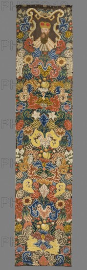 Tapestry embroidered in wool and silk on linen canvas. In the upper part, within a decorative cartouche, is the bust of a man with a mustache, long beard, and wearing a princely mitre. Around it are dense scrolls of large leaves, flowers, and fruit. No border. Vivid colors: brown background, pink, sapphire, green, yellow, and gray ornament. The plant motifs of the nine fabrics comprising this set do not differ from each other. Stylized leaves, flowers, and fruit fill the entire surface of the panel. The composition unfolds on both sides of the axis, defining a symmetrical division of the decoration, similar to the large-pattern silk upholstery fabrics fashionable in the late 17th and early 18th centuries. Minor differences include the inclusion of small animal silhouettes on some panels, enriching the decoration. All fabrics are decorated with the same fruit: plump bunches of black and white grapes, yellow apples, and dark red cherries. Vivid colors, brown background, shaded ornamentat...