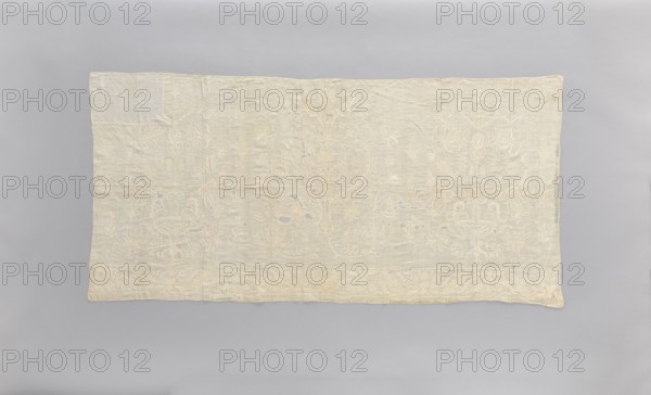 Church napkin made of white linen, covered with white, partially openwork embroidery. In the center is a large stylized flower tree; on the sides are bouquets in vases. The bottom border features a floral tendril motif, punctuated at regular intervals with a candelabra motif. Tall vases and flowers stylized in an Eastern style. Late 18th century, Poland. Fabrics; height 81 cm, width 166 cm., A rectangular textile laid flat against a neutral background, showing an embroidered pattern across its surface; the fabric appears pale cream to off-white with visible lighter embroidered motifs creating a subtle raised design; the background behind the textile is a soft gray; the embroidery includes flowing, symmetrical shapes and floral-like motifs rendered in the same pale cream tone with hints of slightly darker beige in areas of wear or shading, and faint touches of muted bluish-gray showing through in small spots.