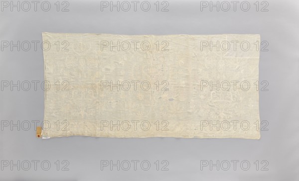 Church napkin made of white linen, covered with white, partially openwork embroidery. In the center is a large stylized flower tree; on the sides are bouquets in vases. The border at the bottom features a floral tendril motif, fastened at regular intervals with a candelabra motif. Tall vases, flowers stylized in the Eastern style. Late 18th century, Poland. Function: church paraments; materials: fabrics; height 81 cm, width 166 cm., A large rectangular piece of fabric with ornate embroidered patterns lies flat against a smooth background; the fabric is a pale cream color with subtle off-white embroidery in floral and curving ornamental motifs that repeat across its surface, and there are a few small areas where the weave appears thin or worn, revealing slightly darker spots; the background is a uniform light gray, and a small tag in tan and a small piece of white material are visible near one corner.