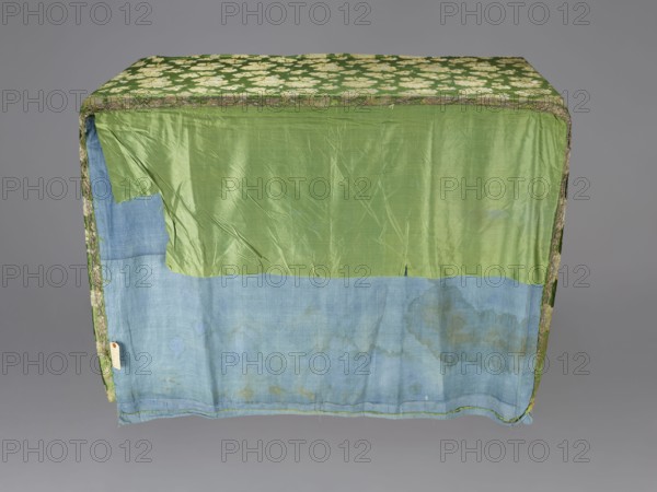 Church napkin, church paraments, evangelical churches furnishings, Sielec Swietokrzyskie Voivodeship, Staszów County, evangelical church Sielec furnishings, decorated with flowers, leaves, and plant ornaments, sacred art, churches and chapels furnishings, 17th century, Italy, fabric, height 120 cm, width 62 cm, A rectangular fabric-covered object photographed against a neutral gray background, showing the front and top surfaces; the top is covered in a green cloth patterned with pale cream floral shapes and edged with a narrow border that includes green and brown tones; the front surface is divided horizontally with an upper section of shiny light green fabric that appears slightly wrinkled and a lower section of faded blue fabric that shows darker stain-like discolored areas and subtle texture variations; the left vertical edge reveals a strip of the patterned green-and-cream fabric and a small hanging tag obscured from readable detail; overall visible colors include green, cream, blu...