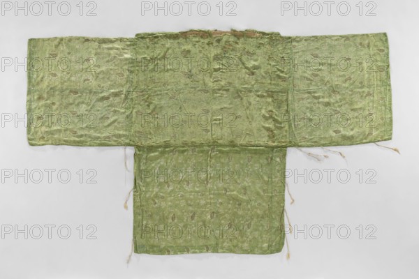 Church napkin; unknown creator; church paraments; Evangelical churches furnishings; Sereje Serejai, Lithuania furnishings; flowers, leaves, plant ornaments; sacred art; churches and chapels furnishings; 17th century, Italy place of origin; fabric; height 180 cm, width 301 cm., A large fabric garment laid flat with a T-shaped form, showing a wide horizontal section and a narrower vertical section extending downward; the fabric appears wrinkled and worn with several frayed edges and loose threads hanging from the lower and side hems. The textile is predominantly light green with areas of slightly darker green shading and a subtle sheen, and it features a repeated floral or leaf-like woven pattern in a paler green that contrasts gently with the background. The surface shows irregular discolorations and small patches where the weave looks thinner or distressed, and the top center edge has a small worn strip with a tan-brown interior visible. The background behind the garment is a smooth, p...