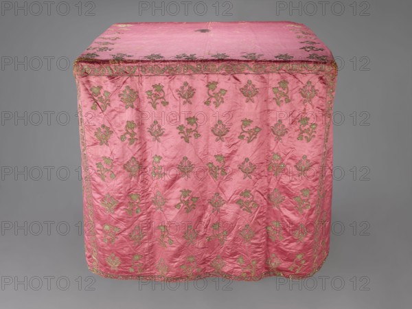 Church cope, liturgical vestment, 17th century, Poland. Made of raspberry satin, embroidered with floral patterns. On the front, branch patterns are arranged in six rows: two branch types—asymmetrical, slightly inclined branches with three flowers, and symmetrical, straight branches with a single flower at the top and two pairs of leaves. Edges decorated with a border of floral and fruit tendrils; lower edges trimmed with gold bobbin lace. Beige linen lining. Height 298 cm, width 201 cm., A rectangular textile draped over a blocky form, predominantly pink with a sheen, featuring a repeating ornate floral motif in a metallic gold-green color across its surface and along its border; the fabric shows visible creases and slight irregularities in texture, with the top panel and sides decorated in the same pattern, and the backdrop is a neutral gray.