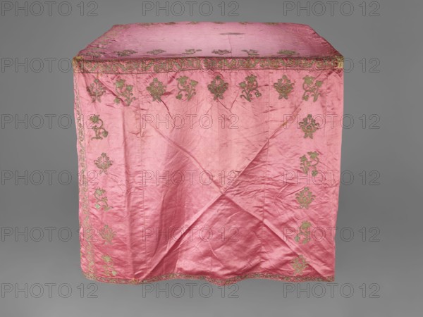 Church cope, gift; liturgical vestment, 17th century, Poland. Made of raspberry satin embroidered with floral patterns. On the front, branch patterns are arranged in six rows: two types — asymmetrical, slightly inclined branches with three flowers, and symmetrical, straight branches with a single flower at the top and two pairs of leaves. Edges decorated with a border of floral and fruit tendril motif; lower edges trimmed with gold bobbin lace. Beige linen lining. Height 298 cm, width 201 cm., A draped pink fabric panel with a slightly wrinkled texture covers a rectangular form, showing a decorative border and repeating ornate motifs embroidered near the edges; the embroidery appears in a metallic gold-green tone contrasting with the pink, and the background around the panel is a neutral gray.