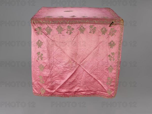 Church cope, liturgical vestment, 17th century, Poland. Made of raspberry satin, embroidered with floral patterns. On the front, branch patterns are arranged in six rows: two types—asymmetrical, slightly inclined branches with three flowers, and symmetrical, straight branches with a single flower at the top and two pairs of leaves. Edges decorated with a border featuring a floral and fruit tendril motif; lower edges trimmed with gold bobbin lace. Beige linen lining. Height 298 cm, width 201 cm., A large pink fabric draped over a rectangular form with visible creases and folds, decorated with a repeating ornate floral motif in a greenish-gold color along the top edge and down the sides, and a decorative border in the same greenish-gold color running around the top and bottom edges; the fabric shows areas of wear and small dark spots, and the background is a uniform gray.