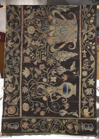 Tapestry. Tapestries. Birds, flowers, coats of arms, fruit; decorative tapestry for manor/home furnishings. Tapestry embroidered on canvas: in the center a basket with flowers, on the sides jugs with flowers and birds. Border with a twig motif. Coats of arms in the corners — top: Jastrzebiec and Korczak; bottom: Poraj Rola and Jelita. Black and brown background; flowers in blue, yellow, green, and red. Embroidery on rare linen canvas in cross stitch with woolen thread; each cross made of two warp threads and two weft threads. 18th century, Poland. Fabrics. Height 158 cm, width 354 cm., A large rectangular textile with a dark brown background fills the image, held at the edges by hands visible at the corners and bottom; the textile is richly embroidered with a repeating floral and foliate pattern in muted colors including beige, cream, pale blue, dusty pink, soft green, and pale yellow. The central field shows an arrangement of stylized flowers, leaves, scrolls, and curving stems, along...