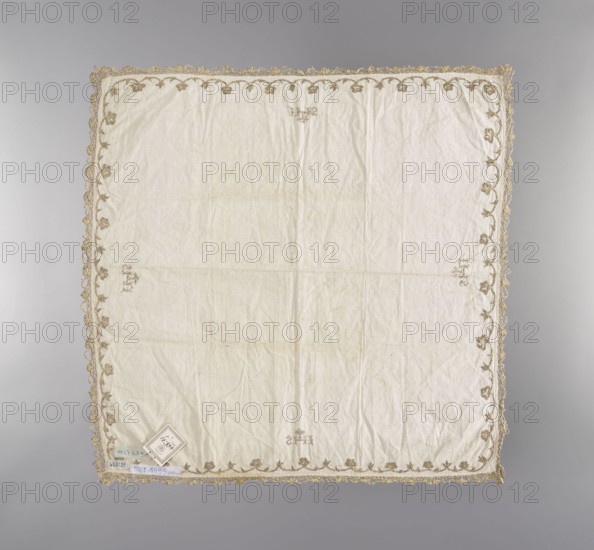 Velum, church paraments, flowers, leaves, plant ornaments, sacred art, churches and chapels furnishings. Velum made of linen batiste embroidered along the edges with gold metal thread in a floral tendril pattern, trimmed with narrow gold bobbin lace. In the center of each edge is the IHS monogram with a cross. 19th century, Germany, liturgical parament, height 42 cm, width 41 cm., A square cloth with a pale off-white center and a decorative border in a muted gold tone, showing a scalloped edge and a repeating floral or leaf motif in the gold thread; the fabric surface is slightly wrinkled with faint shading variations and a few small darker spots; a small diamond-shaped paper or tag lies near one corner on top of the cloth; the background behind the cloth is a smooth gradient of light gray to medium gray.
