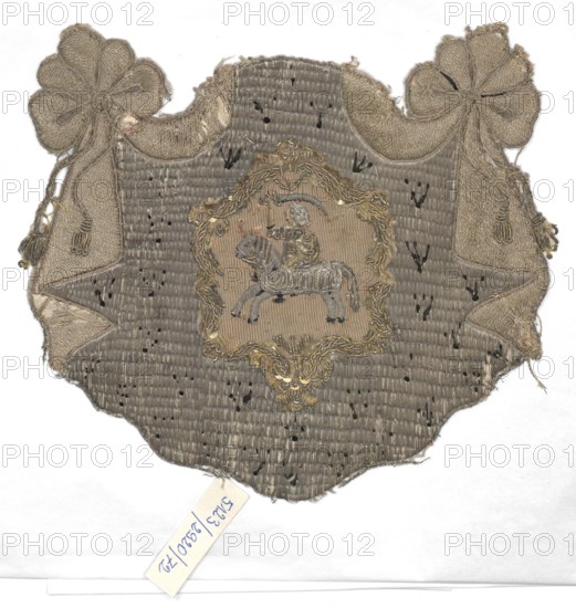 Embroidered coat of arms, embroidery, 18th century, Pogon coat of arms, decorative embroidered fabric, height 16 cm, width 19 cm, provenance unknown, A worn fabric patch in shades of beige, tan, gray, and muted gold is shown against a white background, with frayed edges and a scalloped lower border; the central area features an embroidered framed motif with metallic thread and pale gold embellishments surrounding a stitched depiction of a figure on an animal, both rendered in muted gray and beige tones, and the patch includes decorative bow-like shapes with tassels at the upper left and right in similar beige and tan hues, plus visible small dark spots and scattered areas of wear across the surface.
