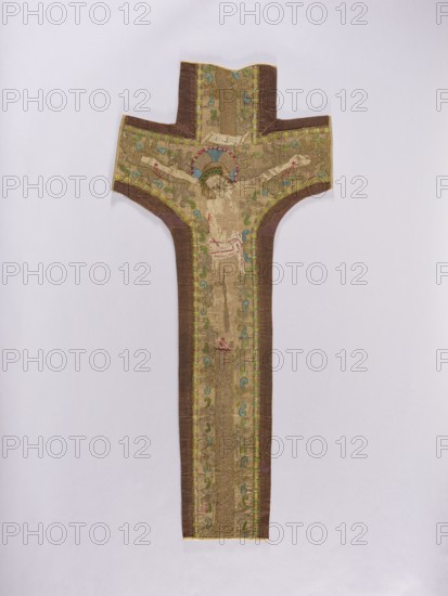 Embroidered Y-shaped chasuble pretext with a depiction of Christ on the cross, surrounded by a stylized plant ornament. Predominant colors: gold, blue, green, and white. Embroidery on silk fabric plain weave with gold metal thread and colored silk threads. Gold cross, surrounded by gold, green, and blue branches. The body of Christ embroidered with white threads that have almost completely fallen out. Metal thread embroidery: laid, held with silk yarn. Silk embroidery: stitches - Cologne, split, flat. 16th century, Poland. Fabrics, length 103 cm, width 46 cm., A long, cross-shaped textile panel displayed against a pale background, with a central vertical shaft and horizontal arms, predominantly in shades of beige and brown with gold-toned embroidery; the central area features an embroidered human figure with outstretched arms, a halo rendered in blue and red above the head, and green decorative motifs flanking the figure; the piece has darker brown borders along the sides and arms, wit...