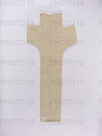 Embroidered Y-shaped chasuble pretext with a depiction of Christ on the cross, surrounded by a stylized plant ornament. Predominant colors: gold, blue, green, and white. Embroidery on silk fabric plain weave with gold metal thread and colored silk threads. Gold cross, surrounded by gold, green, and blue branches. The body of Christ embroidered with white threads that have almost completely fallen out. Metal thread embroidery: laid, held with silk yarn. Silk embroidery stitches: Cologne, split, flat. 16th century, Poland. Fabrics. Length 103 cm, width 46 cm., Iconography: Christ Crucified; function: liturgical vestment church paraments, sacred art; motifs: flowers, leaves, plant ornaments., A long, vertically oriented beige fabric piece with a textured weave is laid flat against a pale lavender-gray background; the fabric widens near the top into a cross-like shape with squared horizontal extensions and a central upward tab, while the lower portion narrows into a long rectangular tail, ...