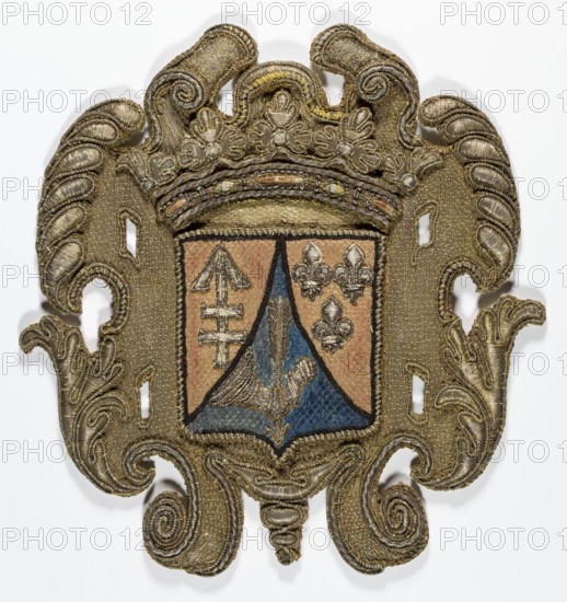 Coat of arms cartouche, embroidery; coat of arms cartouche with a tripartite shield under a crown in the center. Coats of arms: Lis, Kikieryc, unknown most likely the coat of arms of the Sapieha family. 17th century, Poland. Materials: fabrics. Dimensions: height 22 cm, width 21 cm. ., An ornate embroidered shield-shaped patch with an elaborate scrollwork border in metallic gold tones; the central shield area is divided with a triangular blue section and surrounding tan sections, and contains embroidered motifs including a winged element in blue and tan and three fleur-like shapes in metallic tones; the top of the patch features a crown-like decorative element in gold, and the entire piece shows textured stitching, raised metallic threads, and areas of darker and lighter gold, along with visible tan, blue, and muted silver-gray hues.