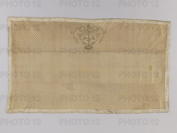 Antependium, antependium; coats of arms; churches and chapels furnishings; carp scale ornament. Made of silver-plated silk fabric with a carp scale motif. At the lower edge in the center is an embroidered cartouche with the Korab coat of arms and a mitre. Above the cartouche is a bishop's hat; at the bottom the cartouche is framed by two branches. Sewn from multiple fabric fragments. Edges trimmed with silver braid. Late 18th century, Sluck possibly fabric origin, Poland. Material: fabric. Height 55 cm, width 97 cm., A rectangular textile laid flat with a repeating small scalloped scale pattern across its surface, bordered by a narrow woven trim. The fabric is primarily shades of gold and beige with slight variations in tone, and there are areas of darker gold and light brown discoloration. Near the top center there is an ornate decorative motif in a muted darker gold. The textile shows faint creases and slight wrinkles and a few subtle spots and stains. The background behind the texti...