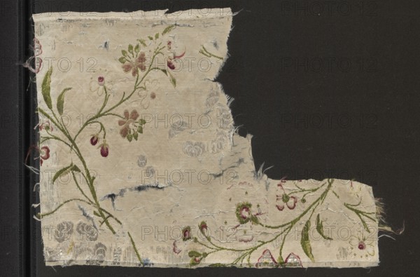 Fragment of white silk grosgrain brocaded with colorful threads and silver stitching; brocade threads include silver stitching, silver metal thread, light celadon, green, grass green, burgundy, apricot, dark pink. Grosgrain background brocaded with small twigs with twining shoots and fancy flowers; drawing includes a rhomboidal mesh net. 18th century, France. Fabric fragment, height 31 cm, width 23.9 cm., A worn rectangular fabric fragment with frayed edges sits against a dark background, showing a faded cream base with a floral embroidered pattern in green, pink, burgundy, and pale brown, including stems, leaves, and blossoms; areas of the fabric have small tears and worn spots revealing lighter and slightly bluish threads, and there are a few scattered darker smudges and thin loose fibers along the edges.