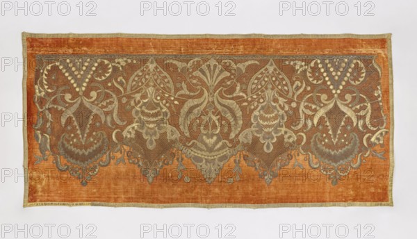 Antependium, antependium; dark yellow velvet with appliquéd decoration of stylized palmettes and flowers in vases in the Louis XIV style; appliqué of gold lace and fragments of gold lamé; edges trimmed with gold galloon; 18th–19th century, Poland; fabric; height 102 cm, width 222 cm., A rectangular textile with a burnt orange background framed by a narrow pale gold border, featuring an ornate symmetrical embroidered band across the upper portion in muted gold and gray tones; the embroidery forms a repeating pattern of stylized floral and foliate motifs with curved, scalloped and lacy shapes, bulbous hanging elements, and flowing tendrils that create mirrored pairs and central vertical designs, with areas of fine mesh-like stitching and solid filled shapes; the overall palette shows burnt orange, pale gold, gray, and subtle brown variations, and the fabric displays slight variations in texture and sheen.