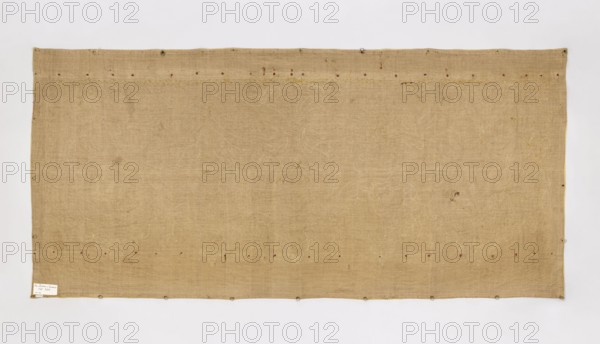 Antependium, Antependium of dark yellow velvet with appliquéd decoration composed of stylized palmettes and flowers in vases in the Louis XIV style. Appliqué of gold lace and fragments of gold lamé. The edges are trimmed with gold galloon. 18th/19th century, Poland. Fabric. Height 102 cm, width 222 cm., A large rectangular textile in a beige-tan color occupies the center, bordered by a pale off-white background; the fabric shows a coarse woven texture with irregularities and faint linear patterns, and there are a few small darker spots and stains scattered across its surface. Several small circular metal rings or eyelets are attached along the lower and upper edges, and a few tiny reddish-brown specks appear near the top and bottom edges. The edges of the textile are slightly uneven and have subtle color variations including pale yellow along parts of the border, and there is a small light-colored rectangular patch near one lower corner.