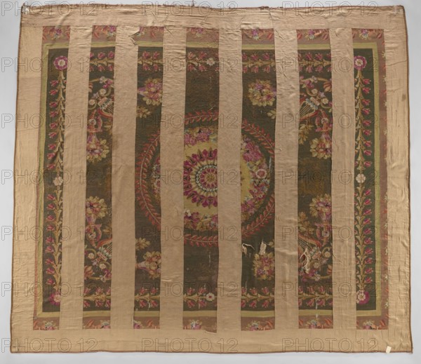 Ornamental tapestry, This late 18th-century Flemish tapestry height 517 cm, width 460 cm — cream, olive, and sand ground with pink, green, and blue ornament — features a symmetrical design: a central rosette surrounded by two floral and leafy wreaths; corner compositions of horns filled with flowers, lyres, and intertwined acanthus leaves; an overall frame of leaves and rosettes; and a border with similar motifs. Materials: woven decorative fabric tapestry., A large square textile with alternating vertical bands of plain tan fabric and ornate patterned panels; the patterned panels show a dark olive background with floral and foliate motifs in shades of pink, rose, red, yellow, green, and cream, including a central circular medallion composed of concentric floral rings; the tan bands and border are a muted beige, and there are visible areas of wear and small holes in some patterned sections; the overall colors visible are tan, olive, pink, rose, red, yellow, green, and cream.