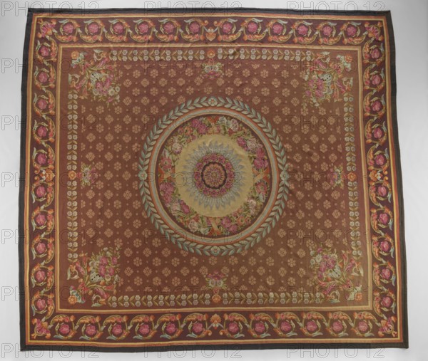 Ornamental tapestry, unknown origin, flowers, floral ornaments, tapestries, weaving, interior furnishings, rosettes, borders, floor tapestry. In the center, a rosette is surrounded by a wreath of flowers, a frame, and a wreath of bay leaves. The entire background of the central field is filled with small rosette motifs; around the edges are simple branches with flowers, and in the corners are compositions of horns with flowers and acanthus leaves. The border features a tendril motif of acanthus leaves and rosettes. Colors: brown-red background, pink, blue, and straw ornament. 1st half of the 19th century, France. Materials: fabrics. Height 512 cm, width 580 cm., A large rectangular textile with a dominant brown background displays a central circular medallion featuring concentric floral and geometric motifs in colors including muted gold, pale blue, pink, green, and beige; the medallion is surrounded by a laurel-like wreath in pale blue and green. The field around the medallion is patt...