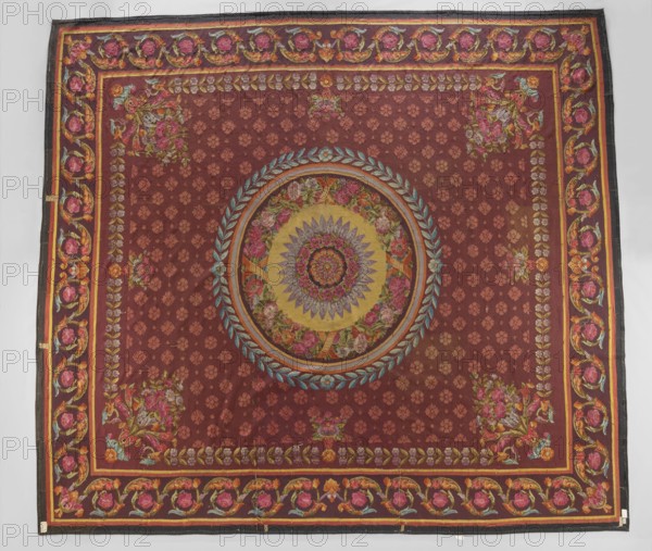 Ornamental tapestry, unknown origin, flowers, floral ornaments, weaving, interior furnishings, rosettes, borders, floor tapestry. In the center a rosette is surrounded by a wreath of flowers, a frame, and a wreath of bay leaves. The entire background of the central field is filled with small rosette motifs; around the edges are simple branches with flowers, and in the corners are compositions of horns with flowers and acanthus leaves. The border features a tendril motif of acanthus leaves and rosettes. Colors: brown-red background, pink, blue, and straw ornament. 1st half of the 19th century, France. Fabrics; height 512 cm, width 580 cm., A large rectangular textile with a deep reddish-brown background featuring a central circular medallion with concentric floral and geometric motifs in colors including gold, yellow, pink, purple, blue, green, and orange, surrounded by a wreath-like ring of blue-green leaves. The field around the medallion shows a repeating small floral pattern in a sl...