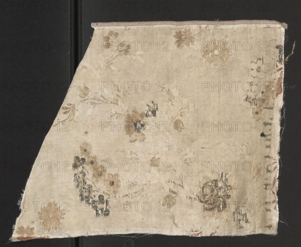 Fragment of fabric, silk fabric fragment with preserved selvedge, patterned floral design on a white background in satin weave with brocaded threads in cream, white fancy, and silver lamellar. 18th century, France. Height 33.5 cm, width 25 cm., A roughly triangular piece of fabric with a worn, light beige background shows a floral woven pattern in muted tan, brown, and dark gray-blue threads; the fabric has frayed edges and uneven borders, with several stylized flower clusters and leafy tendrils scattered across the surface, some areas appearing faded or threadbare, and small loose fibers visible along the margins.