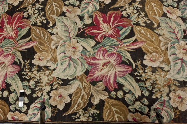 Kilim, Repossession provenance. Flowers; decorative fabrics; modernism style. Manor and home furnishings. Fabrics with plant and floral motifs; weaving. Kilim with motifs of large flowers and leaves filling the entire surface; no border. Colors: black background with light red, green, cream, pink, ash, and brown ornamentation. Late 19th century, Europe. Fabric kilim. Height 725 cm, width 450 cm., A woven textile with a dark background patterned with a floral design in shades of pink, red, cream, beige, tan, light blue, teal, green, and brown; the design includes large stylized blossoms, several broad leaves, and clusters of smaller flowers and buds; along the lower edge two metal tripod legs with black parts are visible and a small rectangular color strip with contrasting panels lies nearby.