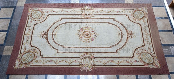 A so-called ceiling tapestry, tapestries, restitution provenance, fabrics, tapestries, knotted monograms, floral motifs, interior furnishings, weaving. In the center, a rosette surrounded by a floral wreath. Surrounded by rectangular frames with rounded shorter sides and floral garlands. In the corners are medallions with a knotted CKJ monogram. The border varies in color with plant patterns imitating the damask technique. Colors: pink, red, light brown, and olive. Early 19th century, France. Fabrics, height 681 cm, width 385 cm., A large rectangular decorative rug lies on a tiled floor. The rug has a wide border in a muted reddish-brown and a central field in a pale cream. Ornate rope-like and floral decorative bands in shades of brown and gold form nested oval and rectangular frames across the rug. Floral bouquets in soft pinks, light yellows, and muted greens appear at several points, including a central floral medallion with a detailed circular motif in brown and gold. Corner medal...